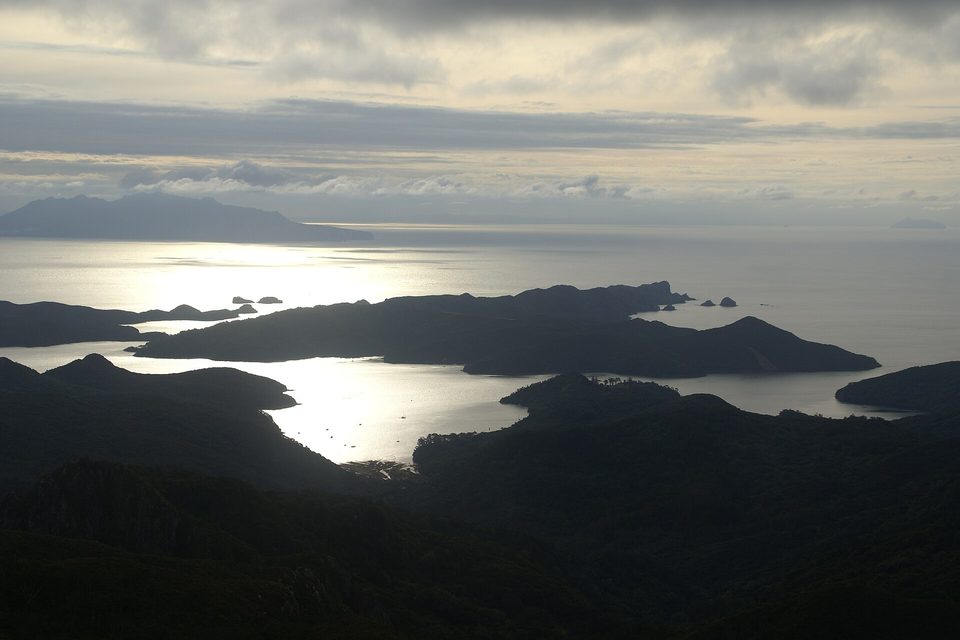 Dark, layered island silhouettes sit in silver evening water, with a long, low island on the far horizon under banded cloud
