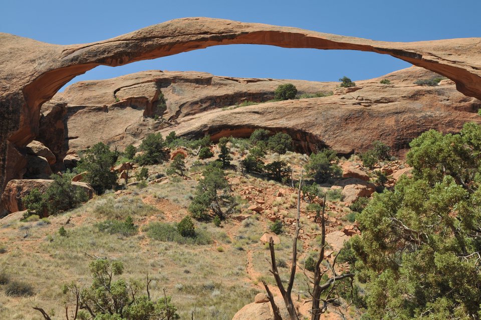 A long, very thin arch of orange sandstone spanning a clear blue sky above a slope of red boulders, grass and dark green junipers
