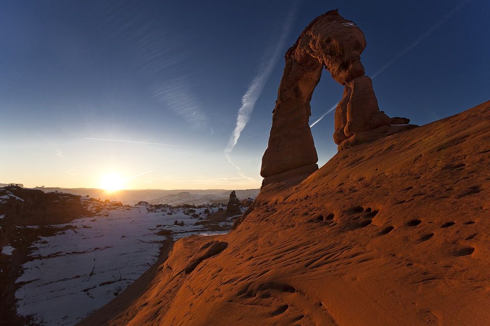 A tall red sandstone arch seen from below against a deep blue sky with two pale contrails, the low sun near the horizon at the left and orange slickrock in the foreground