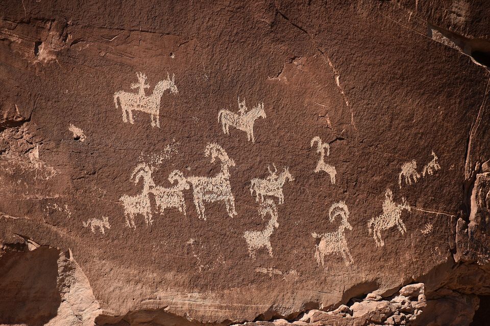 A panel of pale figures pecked into dark brown rock: horses with riders and several bighorn sheep