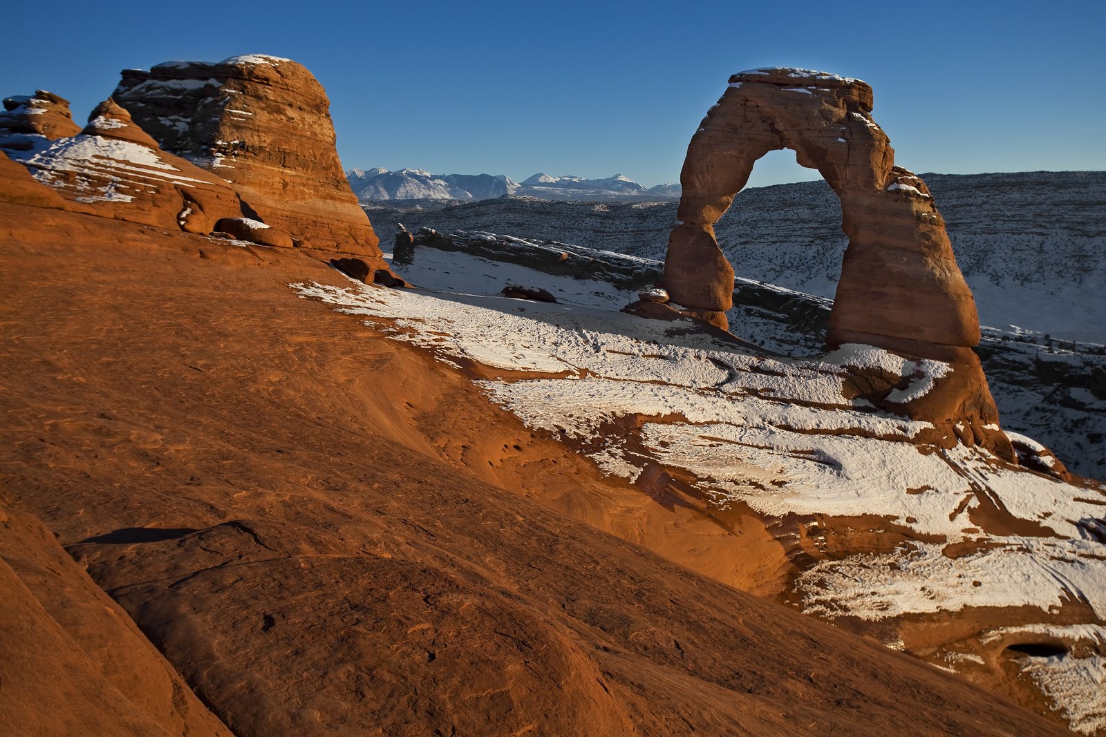 A tall red sandstone arch on the right rim of a rock bowl streaked with snow, a line of snow-covered mountains far off under a deep blue sky, red-orange rock sloping at the left