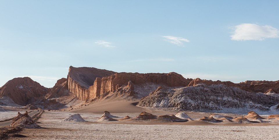 A tilted ridge of layered rock rising above a flat floor of pale salt crust and low mounds under a blue sky