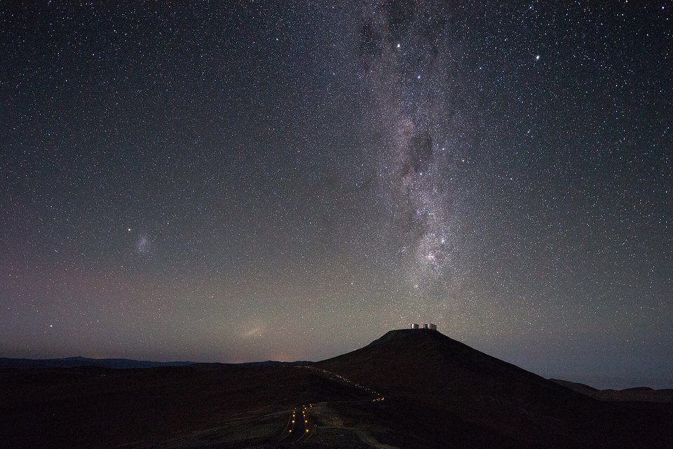 The Milky Way rising in a dense starfield over a dark hill topped with the domes of a telescope observatory