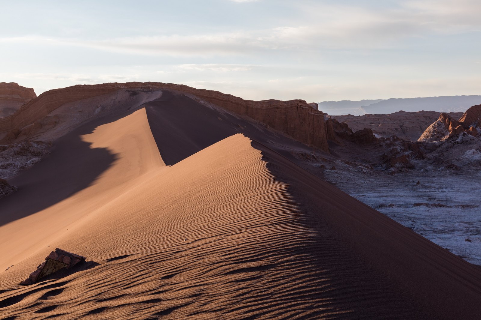 A knife-edged sand dune in evening side light above an eroded valley with pale salt crust and reddish cliffs beyond