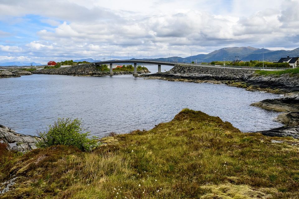 A low concrete bridge on slim piers crossing a calm grey-blue sound, moss and cotton-grass on the rocks in front, red and white houses and mountains beyond