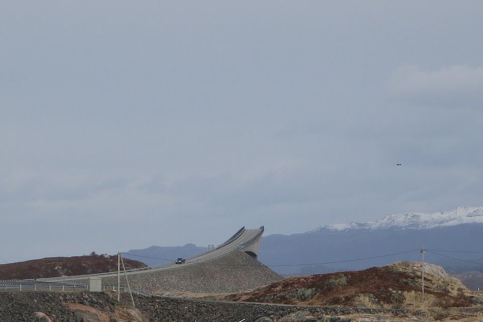 A concrete road bridge climbing steeply and seeming to end in mid-air above a rocky causeway, snowy mountains behind