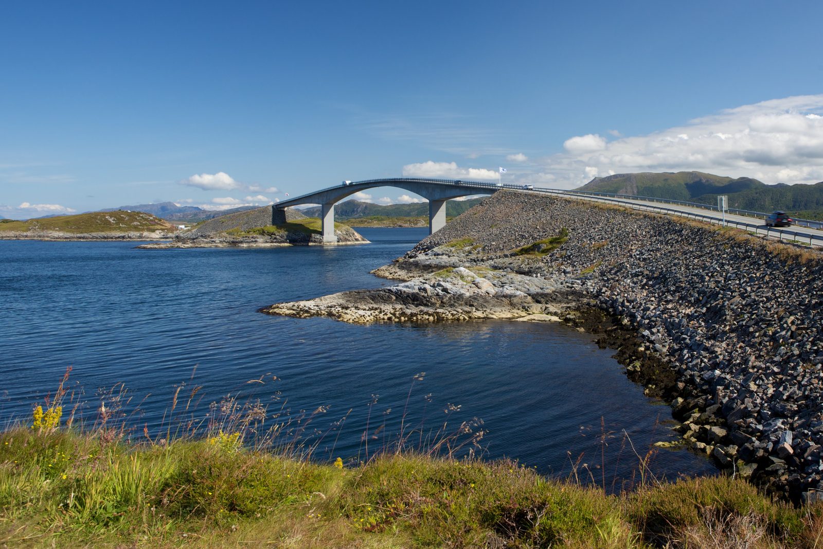 Storseisundet Bridge arching over a calm blue sound from a rocky islet, a stone causeway curving in from the right, green islands and low mountains under a summer sky