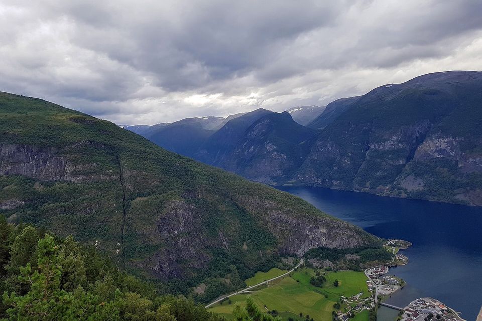 A deep blue fjord between steep green and grey mountainsides, a village and fields on the shore far below, heavy grey cloud above