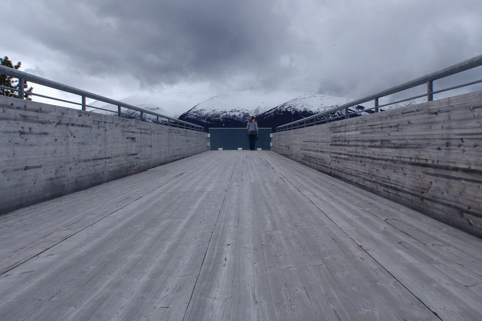 A long pale-timber walkway with steel rails running toward a glass end panel, snowy mountains under dark cloud beyond, one figure standing at the far end seen from behind