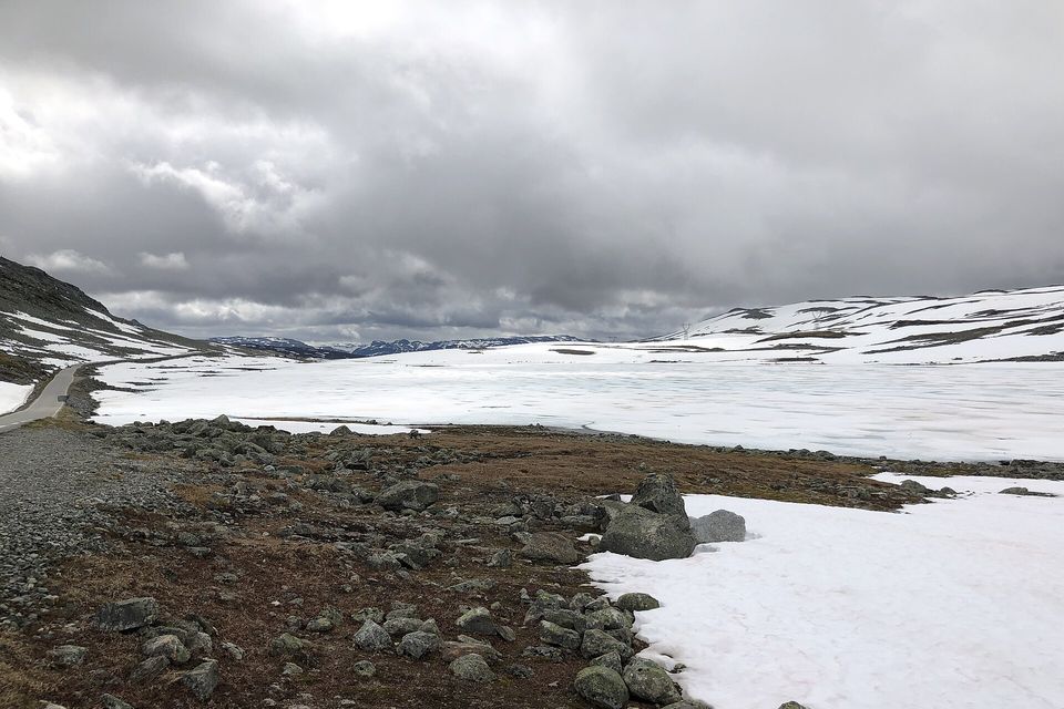 A wide frozen lake covered in grey-white ice and snow, a narrow road running along its left shore, mossy boulders and a snowbank in the foreground under low cloud