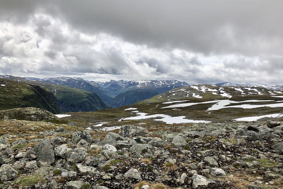 A bare plateau of grey boulders and moss, snow patches spread across the slopes and snowy mountains under heavy cloud