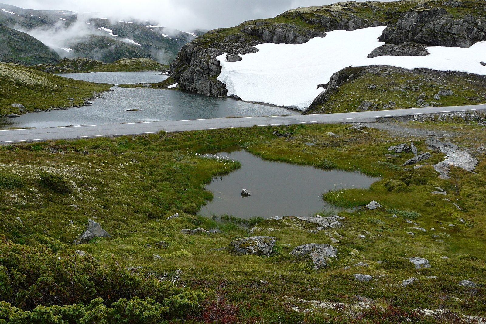 The Snow Road crossing a high, bare plateau between a black tarn and a bank of old snow under grey crags, mist on the mountains behind and heather and lichen in the foreground
