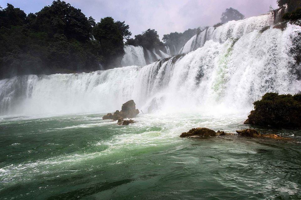 White water pours over a broad ledge into a churning green pool, with rocks in the foreground