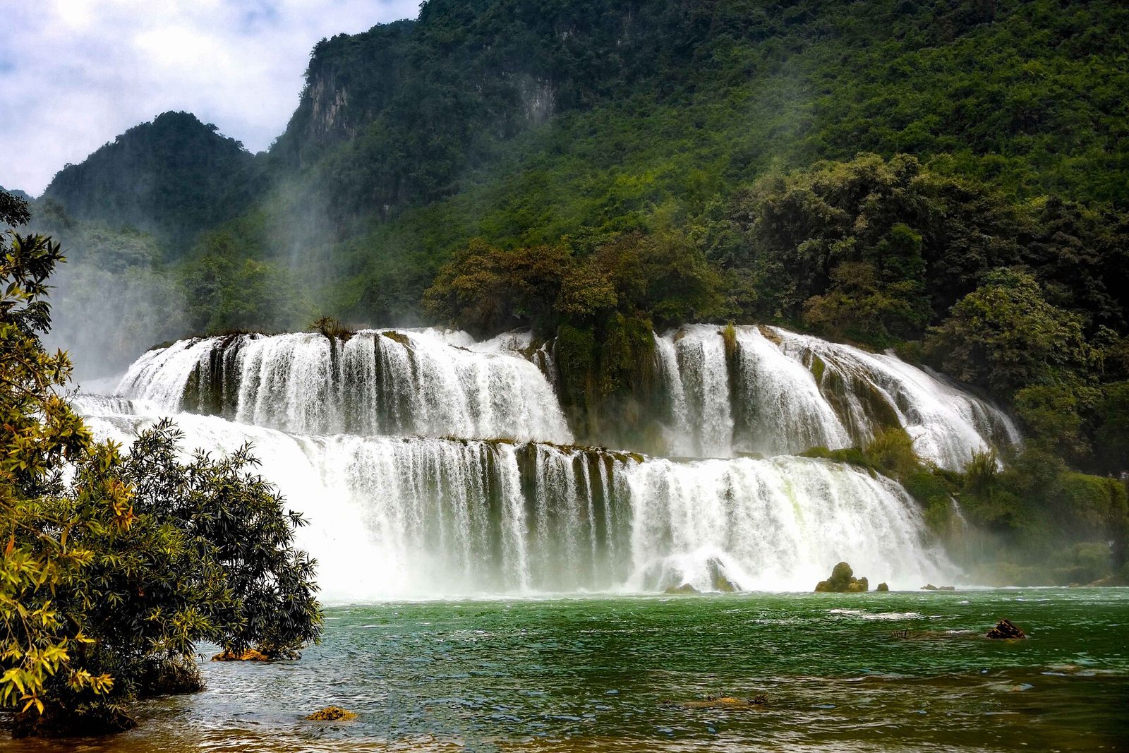A two-tier curtain of white water falls into a jade pool, with spray drifting up a jungle-covered karst wall