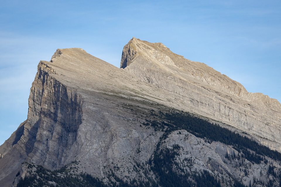 The sloping grey rock face of a mountain peak against a pale blue sky