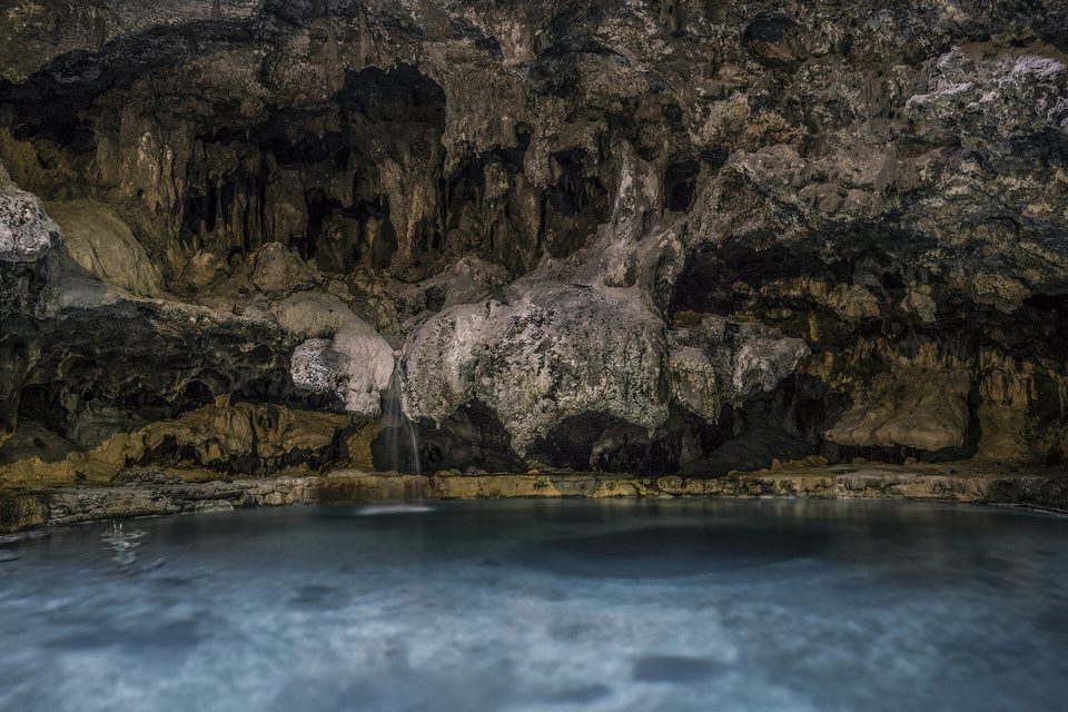 A dark limestone cave with a shallow pool of still, pale water at its floor