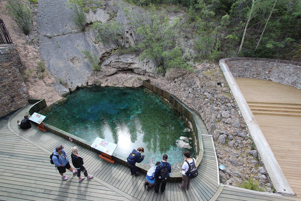 A clear turquoise spring pool below a pale rock wall, visitors looking in from a wooden boardwalk