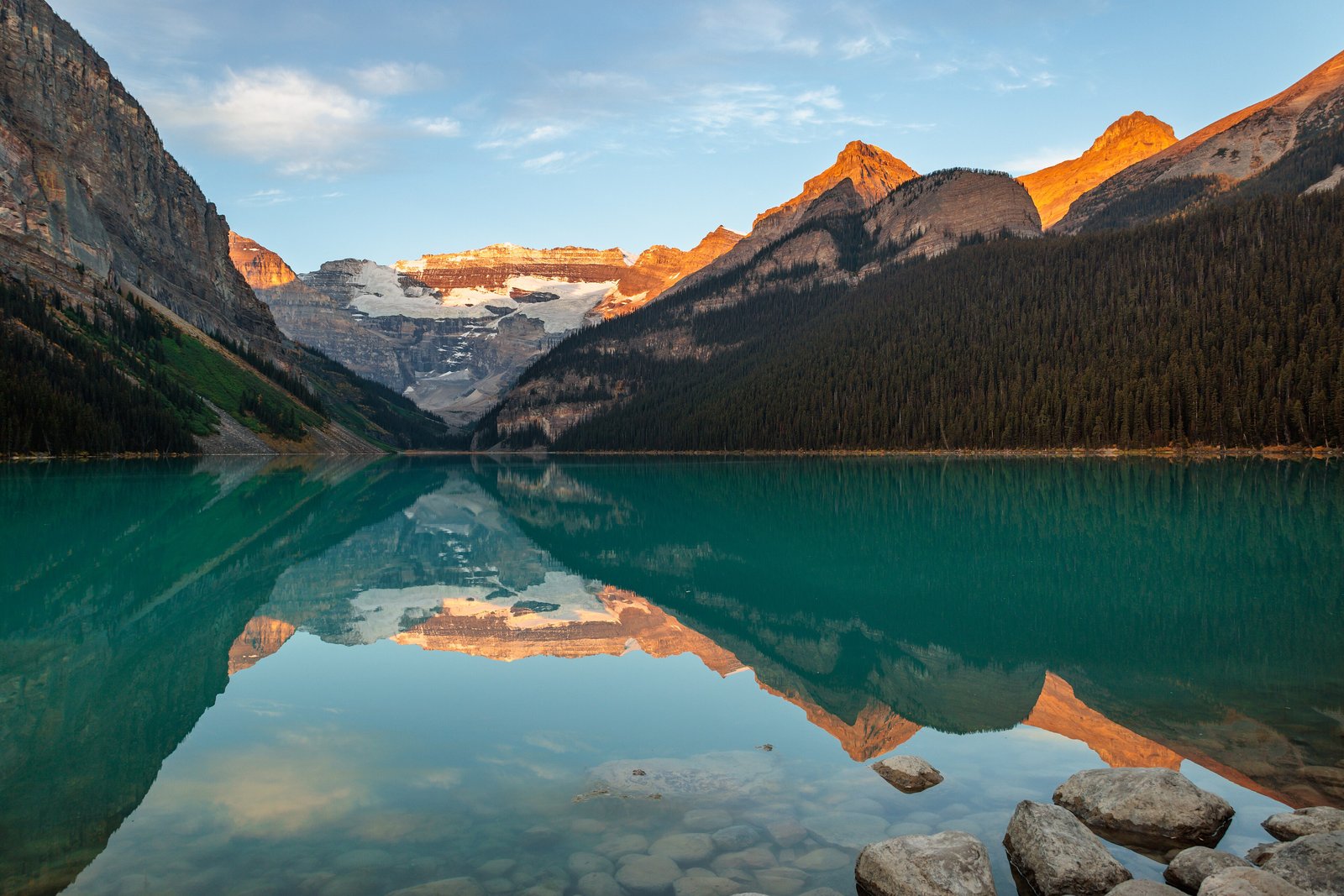 A turquoise mountain lake at dawn, orange light on the far ridge and snowy peaks, dark spruce forest on the right and rocks in the foreground