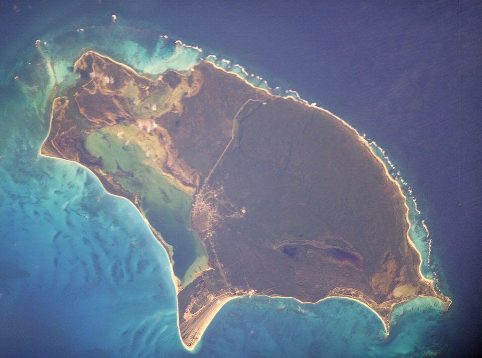 A flat, rounded island seen from orbit, a long pale beach and a shallow green lagoon along its west side and a white reef line on the east