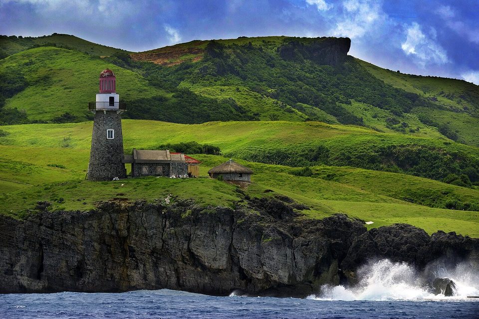 A stone lighthouse with a red lantern stands beside a low building and a round thatched hut on green slopes above dark cliffs and breaking surf