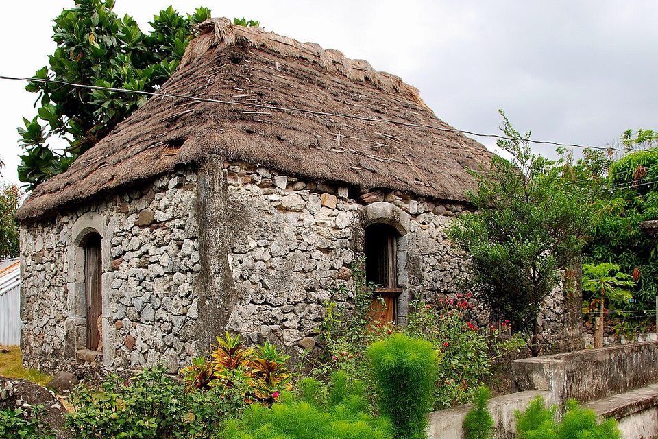 A small house of pale, rough-cut stone with arched doorways under a steep, shaggy thatched roof, framed by garden plants
