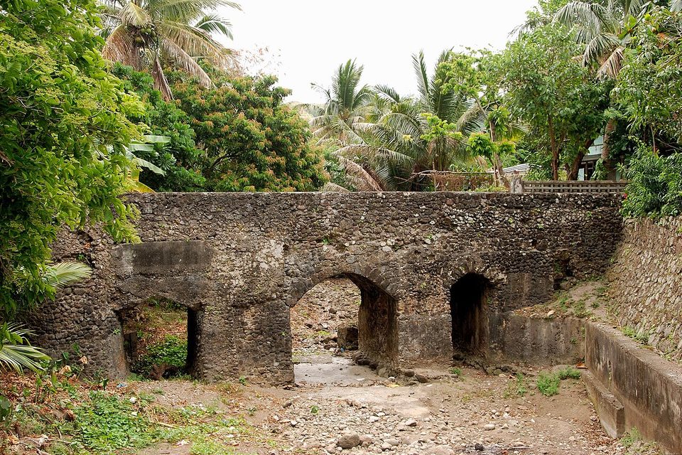 A low stone bridge with three arches spans a dry, stony streambed among palms and green trees