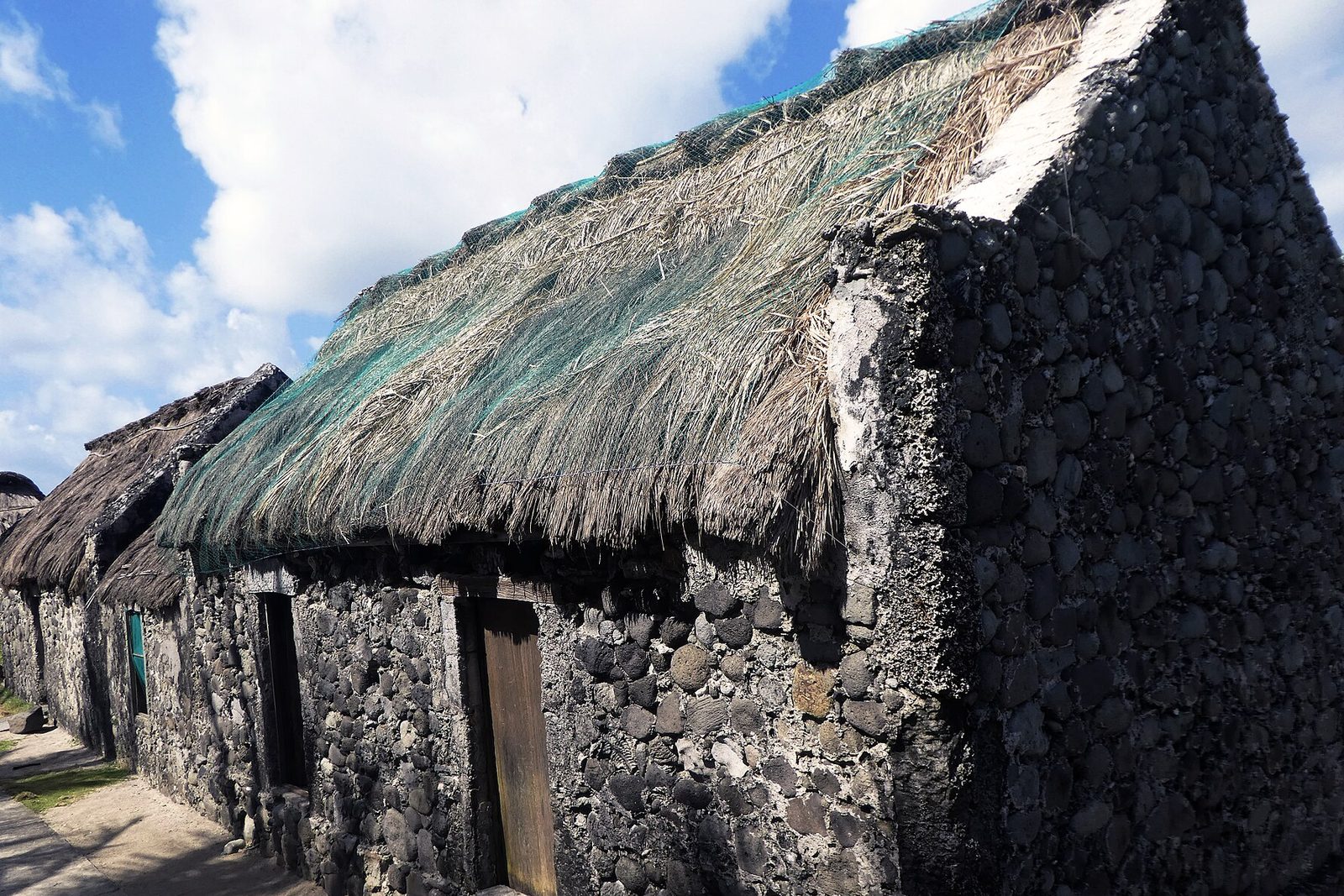 A row of low stone houses of dark rounded stones in pale mortar under thick thatch, beneath a blue sky with cumulus