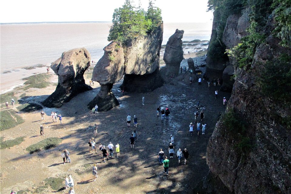 The same sandstone stacks seen from above at low tide, their narrow bases exposed, with dozens of small figures walking on the mud below