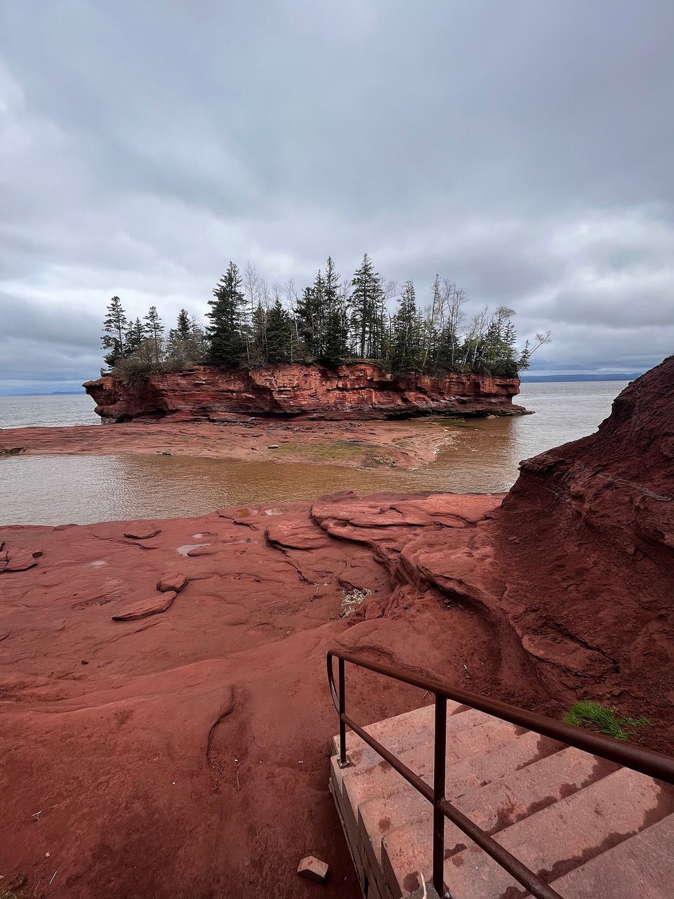 Stone steps with a metal handrail leading down onto a floor of smooth red rock, a tree-topped rock island and brown water beyond