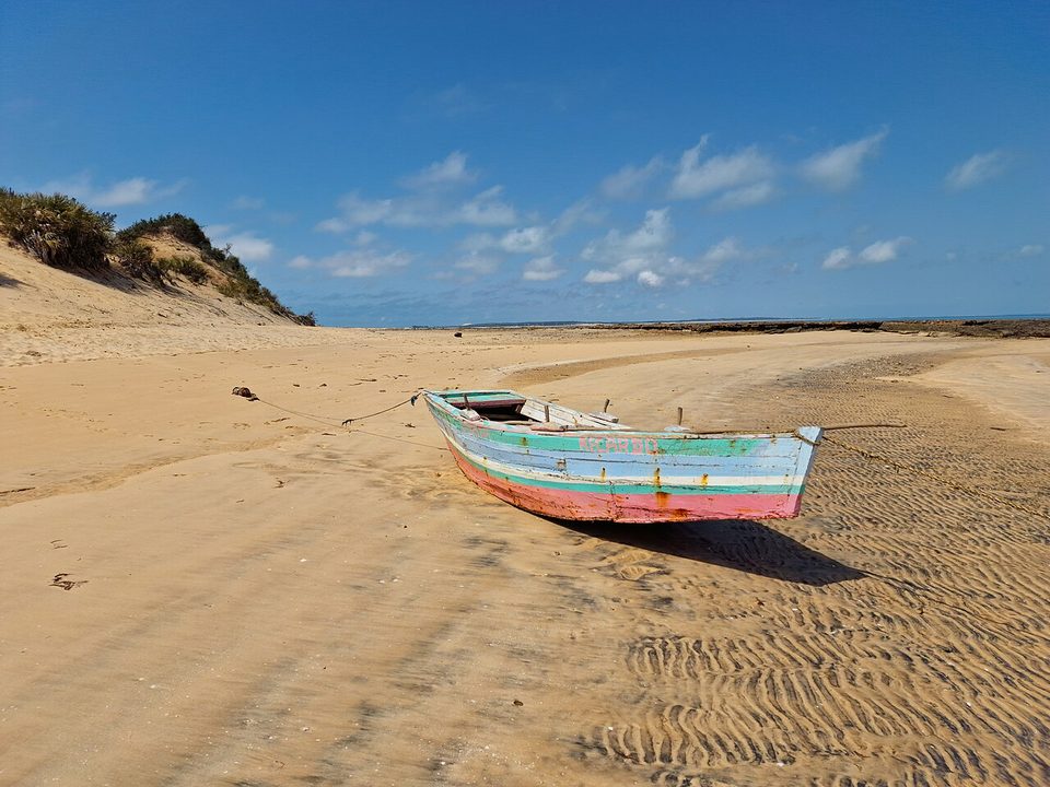 A blue, green and pink painted wooden boat on rippled sand with a dune behind