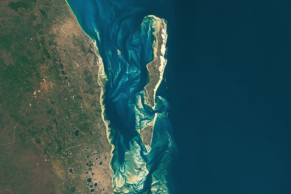 A satellite view of swirling turquoise shoals and sandbars between a long island and the mainland coast