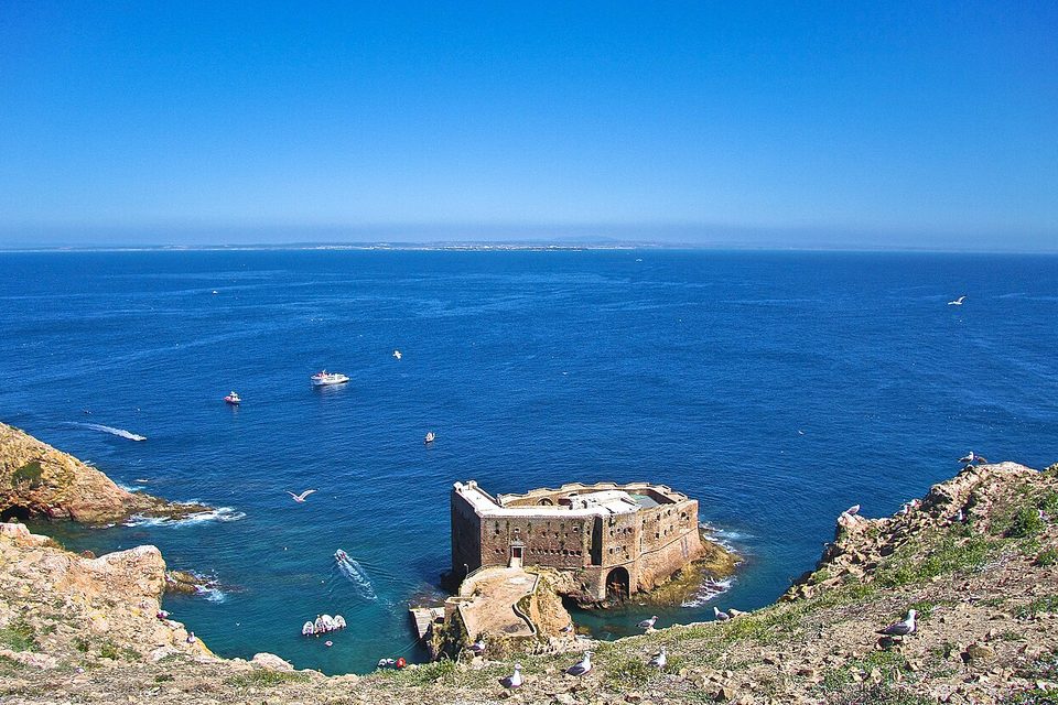 A stone fort on a rock in a deep-blue cove, seen from a gull-dotted clifftop, with a thin line of coast far across the sea