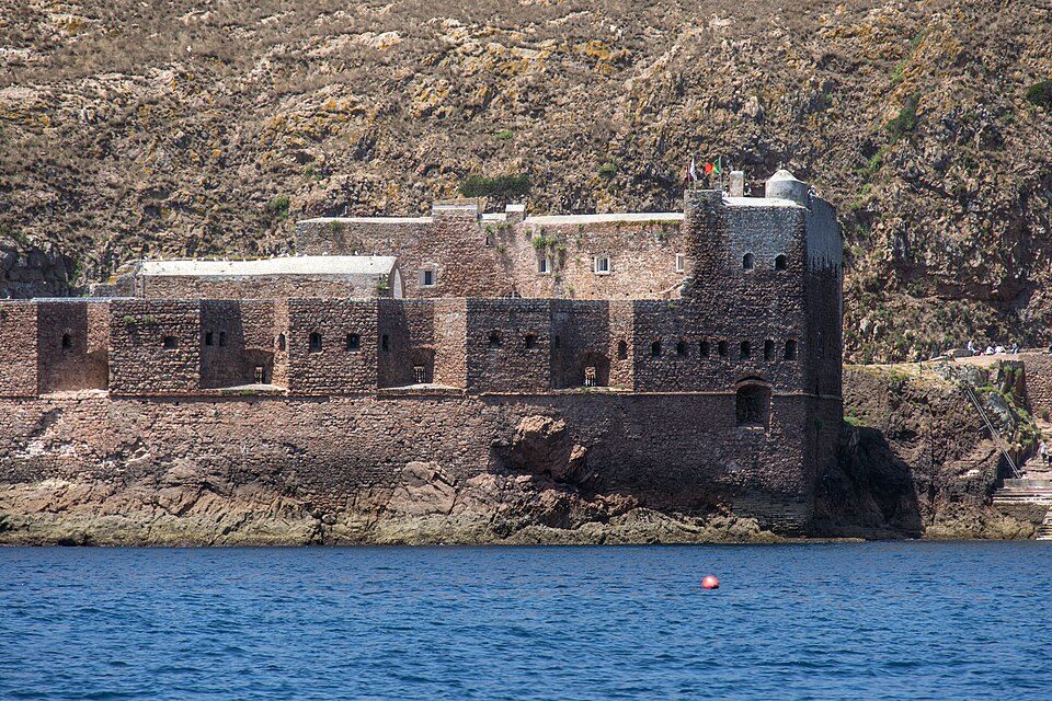 Massive walls of rust-brown stone with small square windows rise from a rock ledge above blue water, a scrubby hillside behind