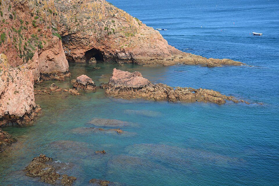 Rose-coloured granite cliffs with a dark sea cave drop into clear turquoise shallows and rock reefs