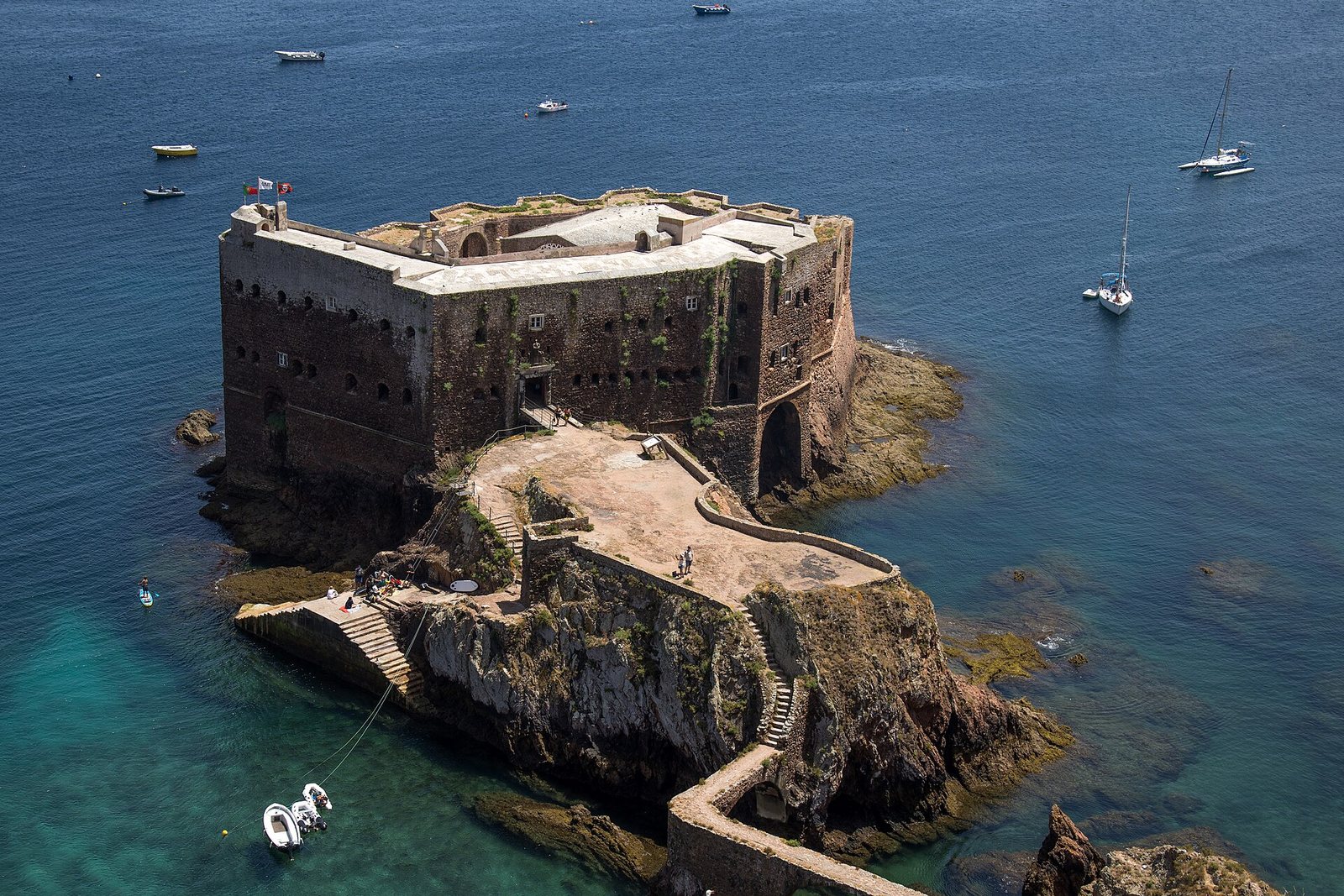 A dark stone fort sits on its own rock islet beside a walled ramp, with jade water at its foot and deep-blue sea beyond
