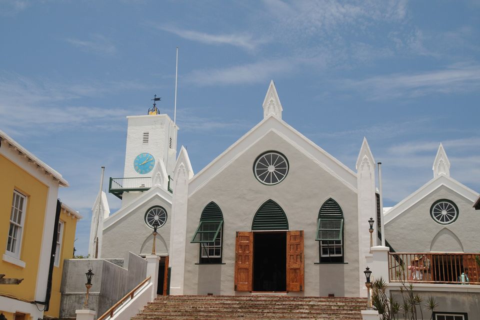 A whitewashed stone church with a square clock tower, green louvred windows and a flight of brick steps, a yellow house at left