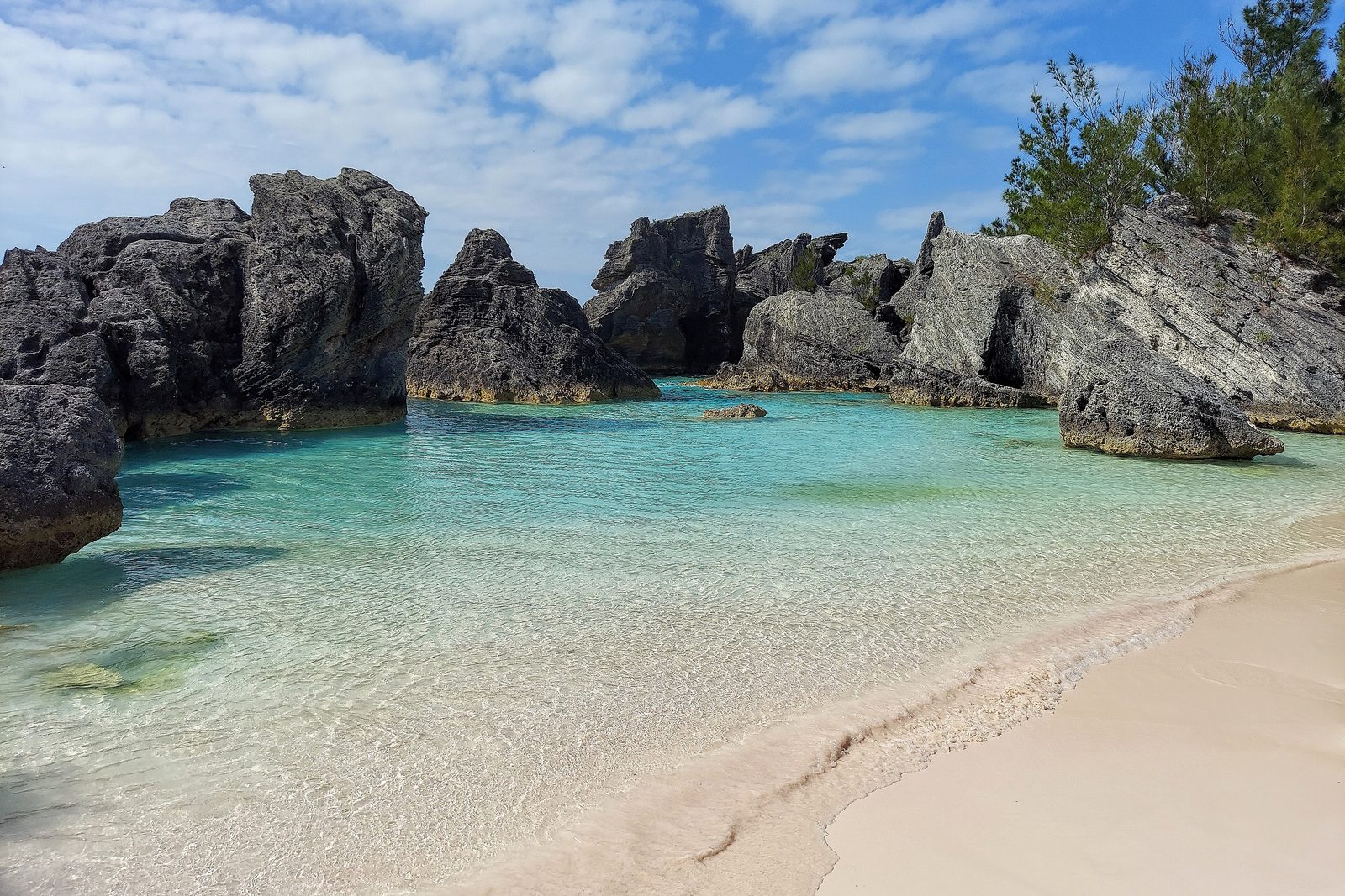 A small cove of faintly pink sand and clear turquoise water between dark eroded limestone stacks under broken cloud