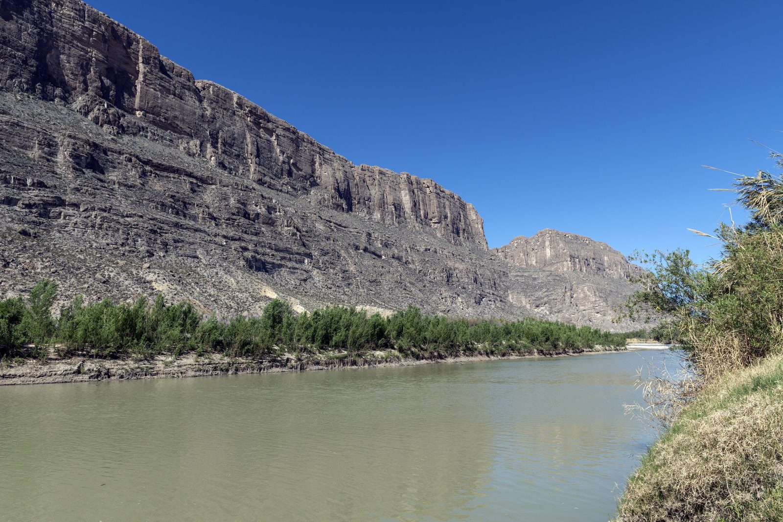 A calm green river curving between a high limestone cliff on the left and a brushy bank on the right, under a blue sky