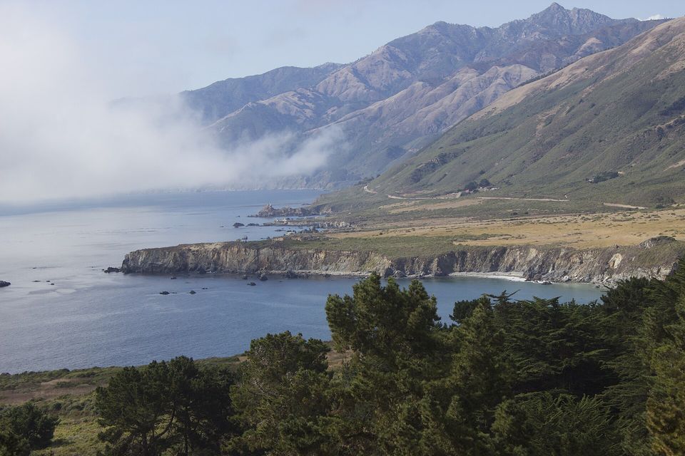 A thin road winding along the foot of steep brown mountains above a headland and a calm bay, fog spilling across the water at the left and cypress trees in the foreground