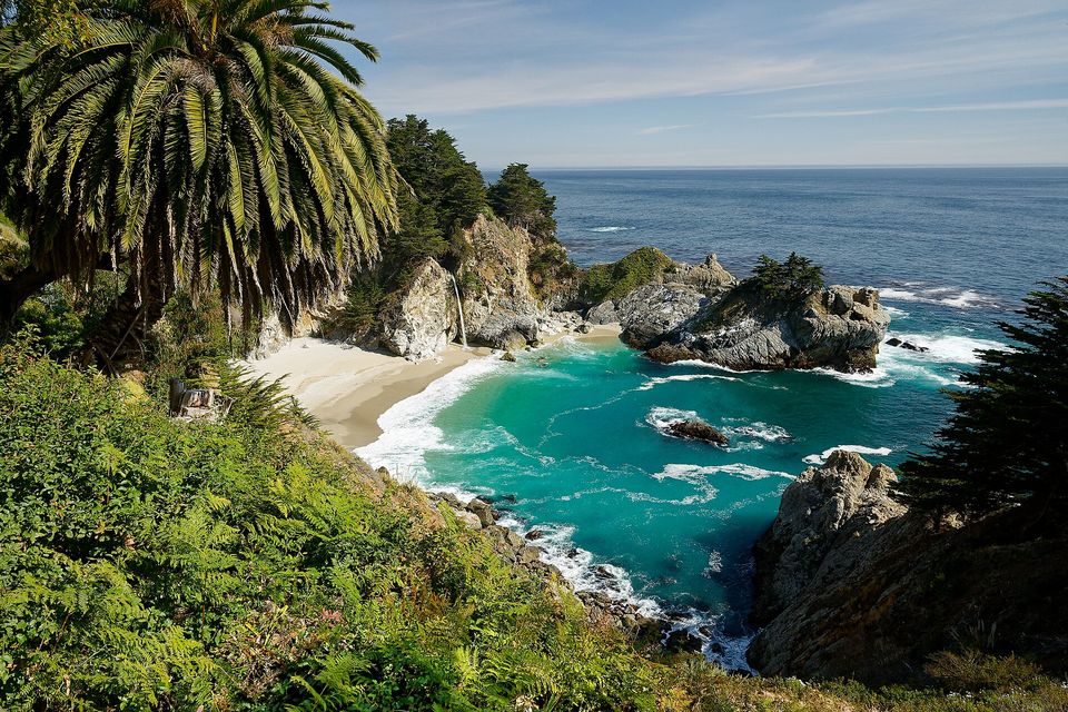 A slender waterfall falling from a wooded cliff onto a pale sandy cove of turquoise water, a palm and ferns in the foreground and the open Pacific beyond