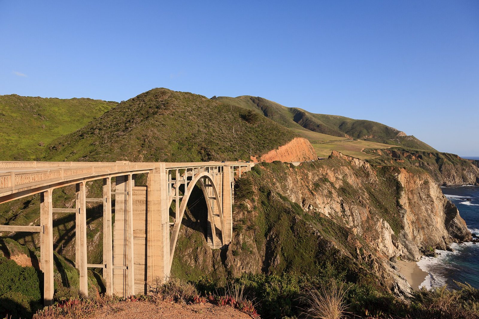 The concrete arch of Bixby Creek Bridge spanning a deep green canyon in low evening sun, the bridge deck running in from the left, sea cliffs and surf at the right under a clear blue sky