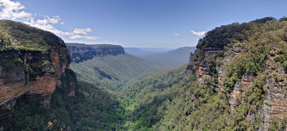 A wide forested gorge between two orange sandstone cliffs, opening onto a distant plateau under a blue sky