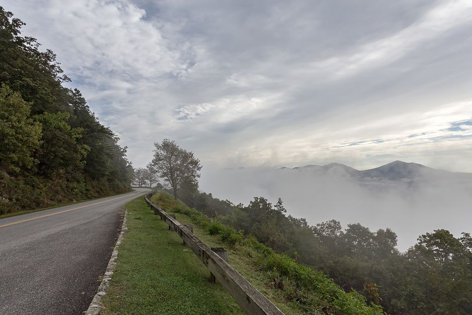 A two-lane road with a wooden guardrail running along a wooded ridge, white cloud pooled in the valleys and dark peaks rising above it