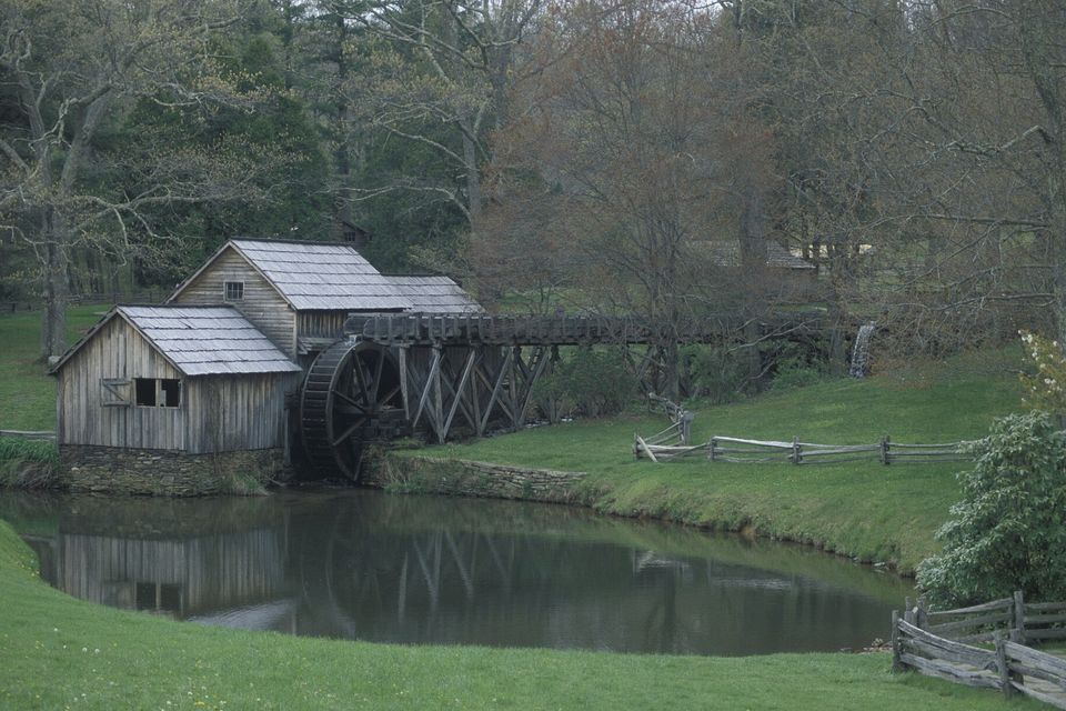 A weathered grey timber gristmill with a wooden water wheel and flume beside a still pond, split-rail fences and spring trees around it