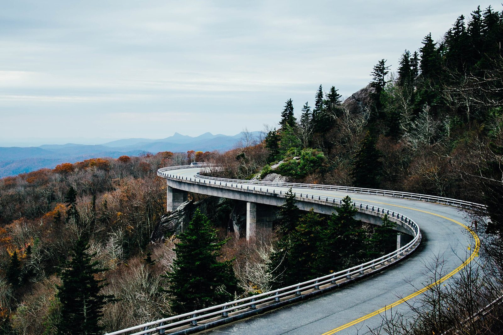 The Linn Cove Viaduct curving around the flank of Grandfather Mountain on slim piers, late-autumn forest in rust and grey, blue ridges fading to the horizon under soft cloud