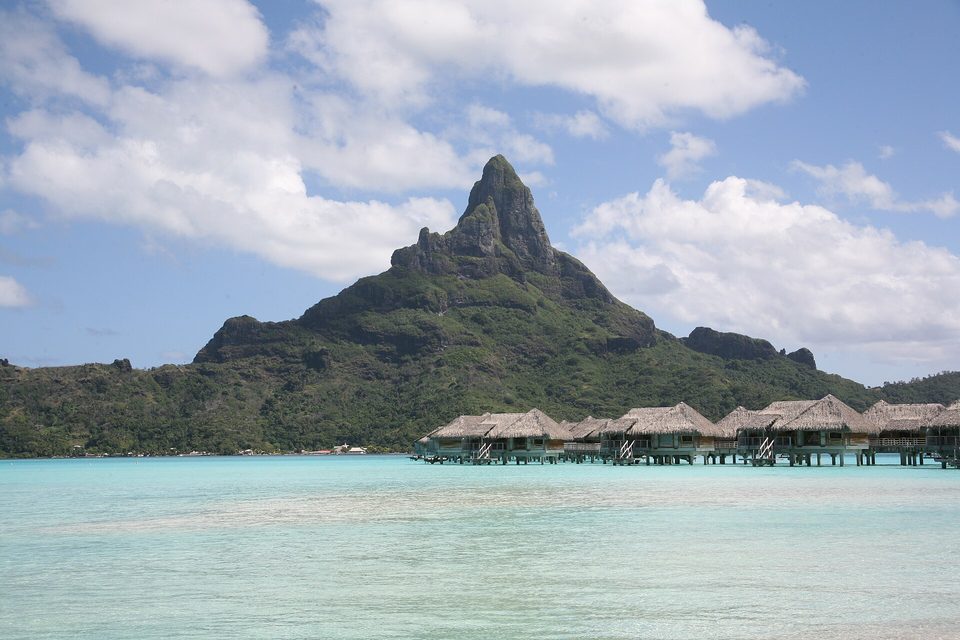 A dark green volcanic peak with a sharp summit above a pale turquoise lagoon, a row of thatched overwater bungalows at the right, white clouds