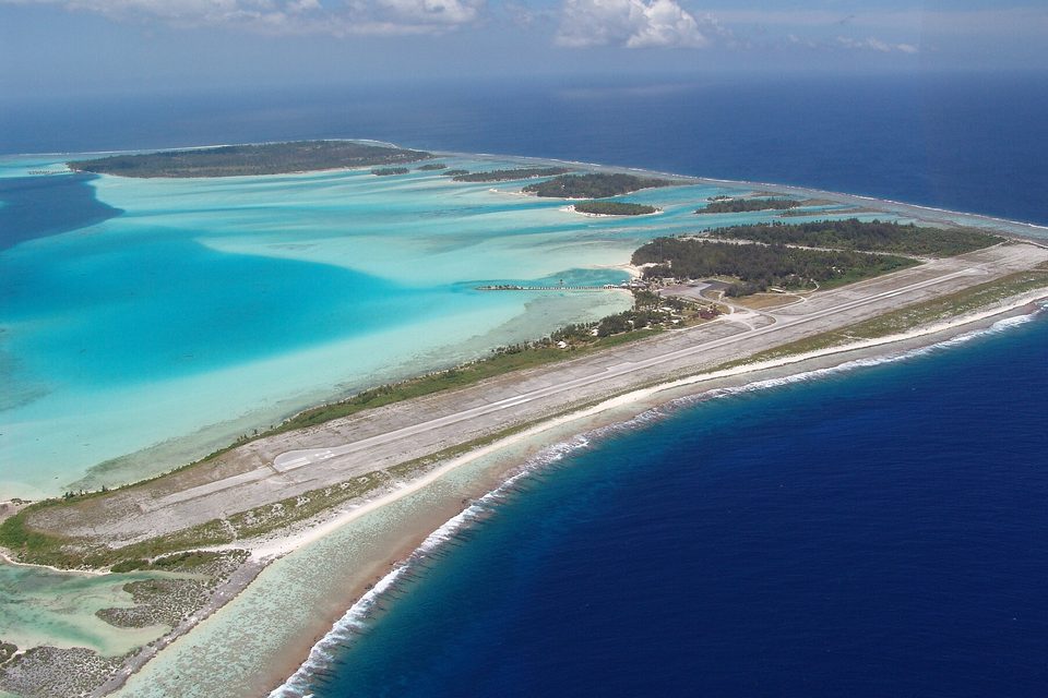 An aerial view of a long runway on a narrow reef islet between a pale turquoise lagoon and the deep blue ocean, a line of surf on the reef