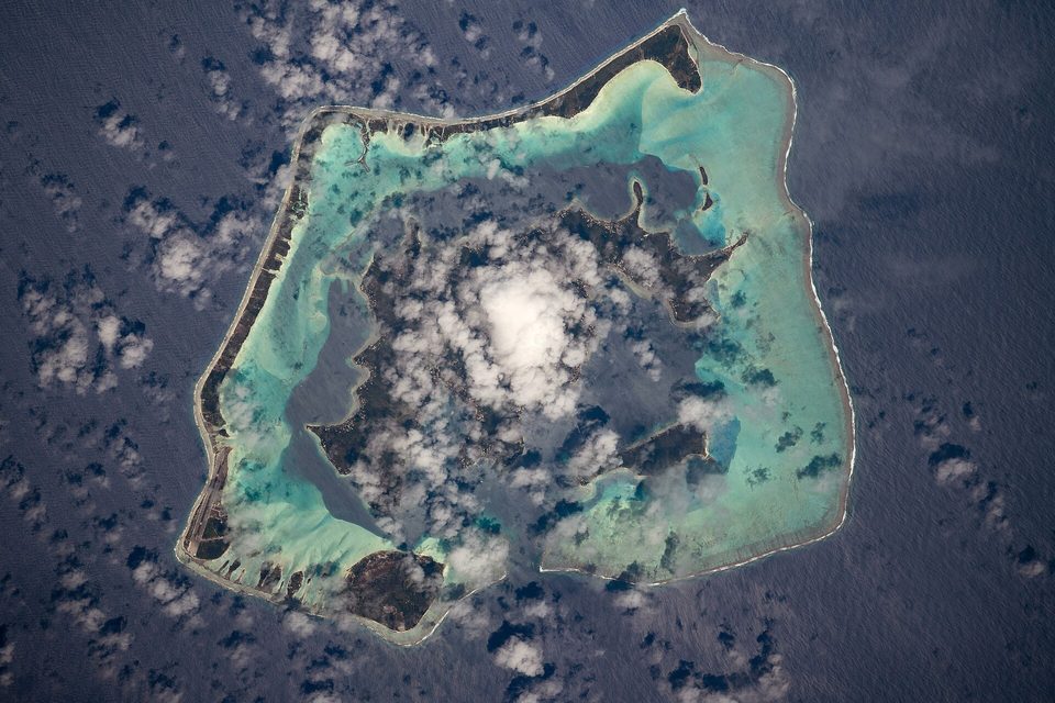 A view from orbit of a ring-shaped reef around a green island and pale lagoon, scattered clouds and their shadows on dark blue sea