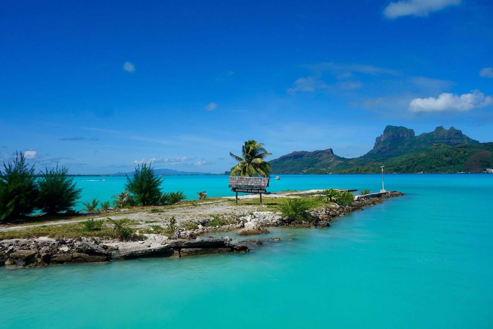 A rocky jetty with a palm and a small wooden airport sign reaching into a vivid turquoise lagoon, a green volcanic peak across the water under a deep blue sky