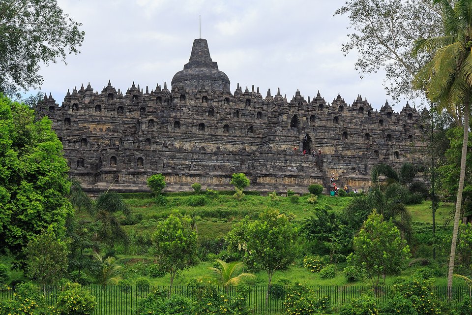 A broad stepped stone monument crowned by a central bell-shaped stupa, seen across green gardens and trees under a pale sky